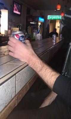 a man holding a can of PBR at Turtle & Inky's while Rocky V plays on the TV and the bartender sits next to a "Carpenter" sign.